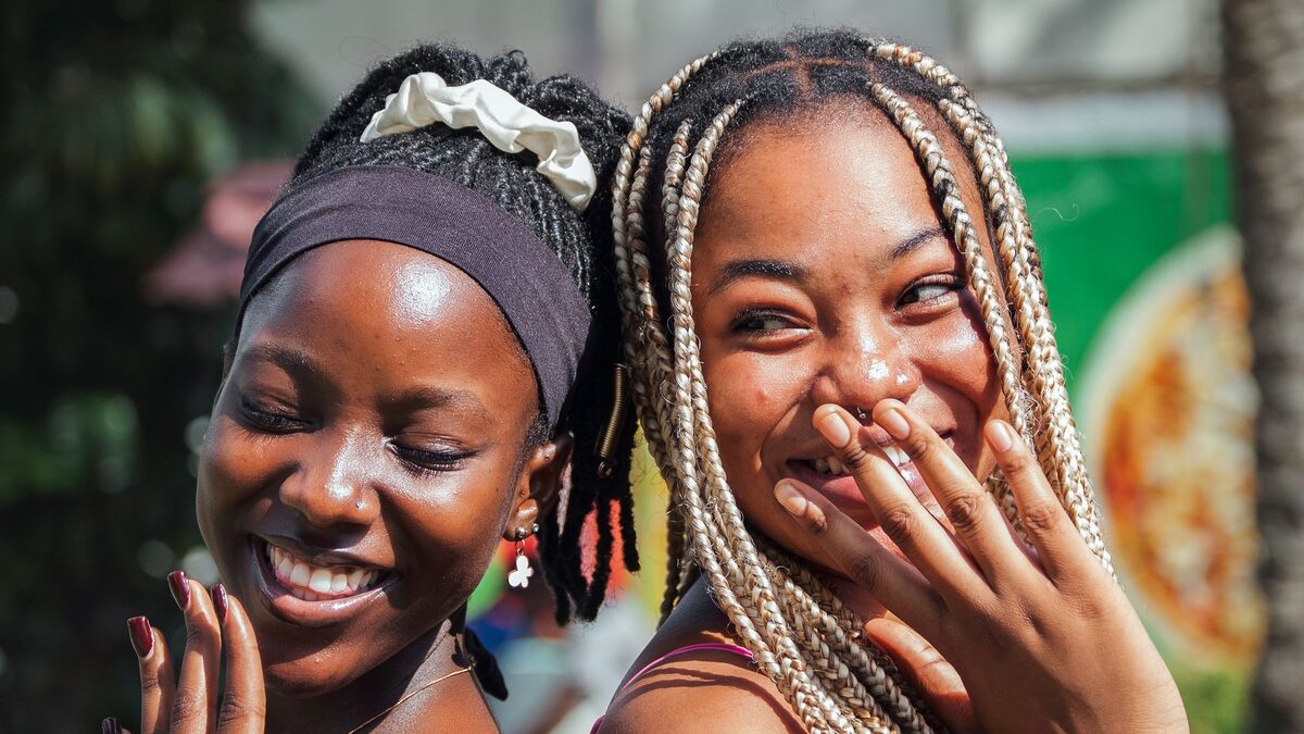 Two young women with braided hair laugh together outdoors in bright sunlight, one covering her mouth with her hand.